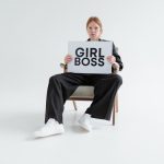 A woman in a black suit holds a 'Girl Boss' sign, exuding confidence on a plain background.