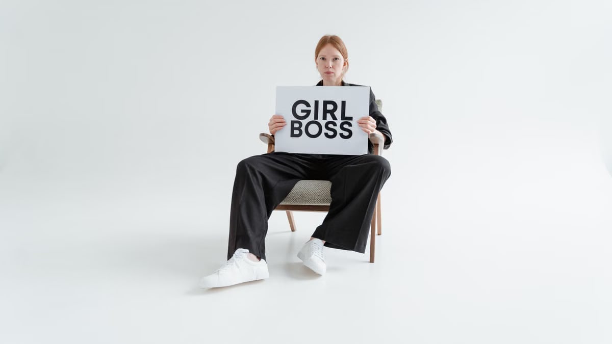A woman in a black suit holds a 'Girl Boss' sign, exuding confidence on a plain background.